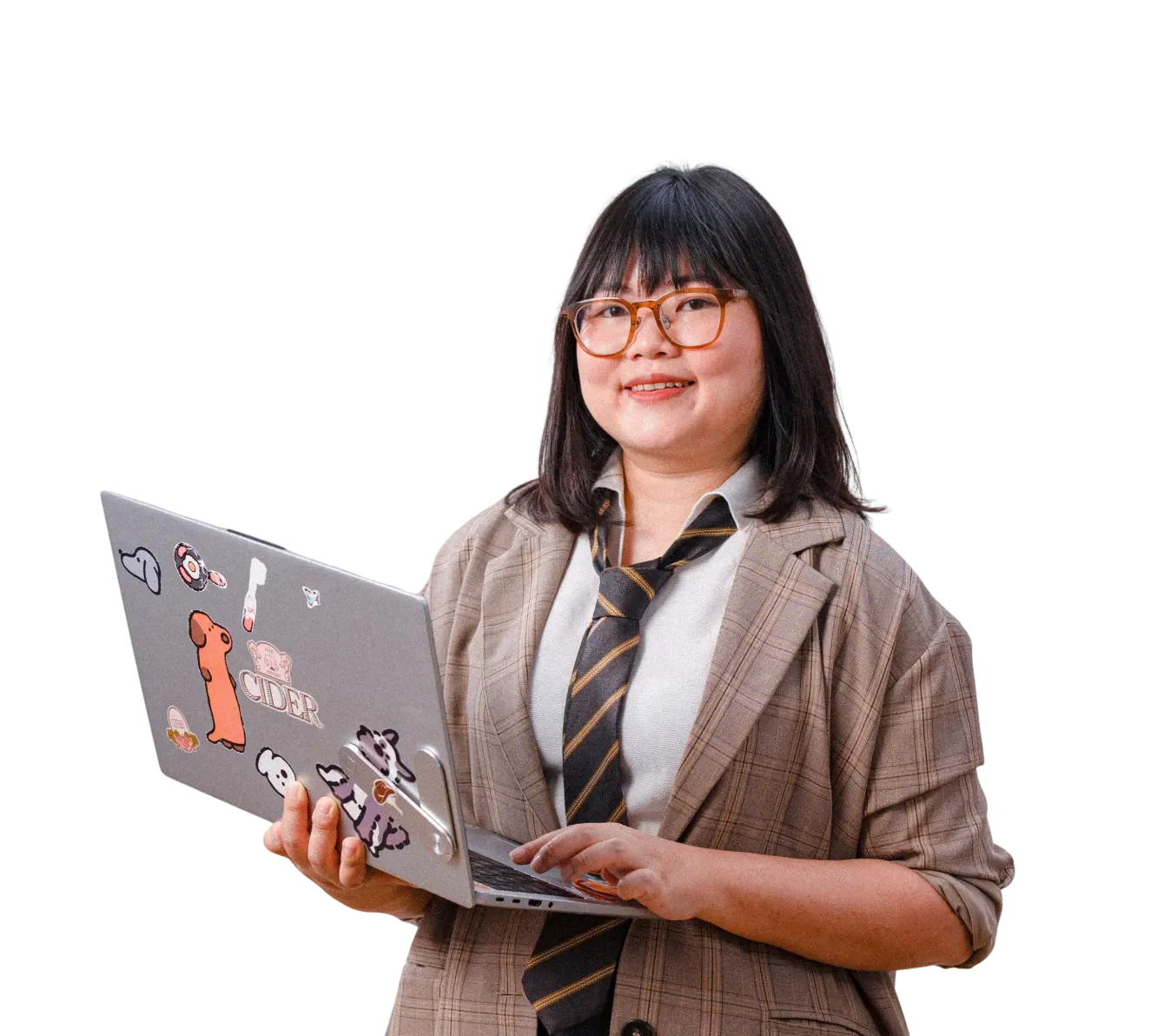 A woman with glasses and a plaid blazer holding a laptop decorated with stickers, smiling while standing.