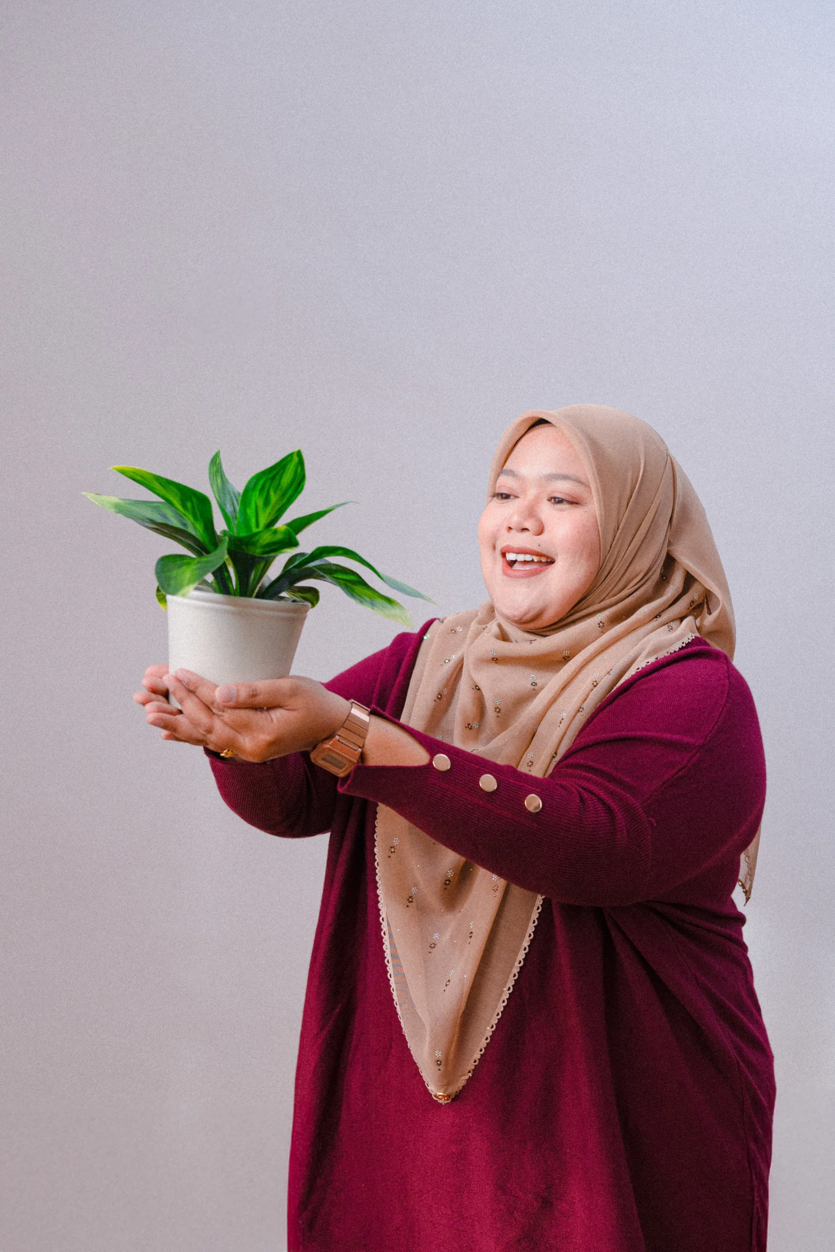A woman in a maroon dress holding a potted plant, smiling while presenting it.