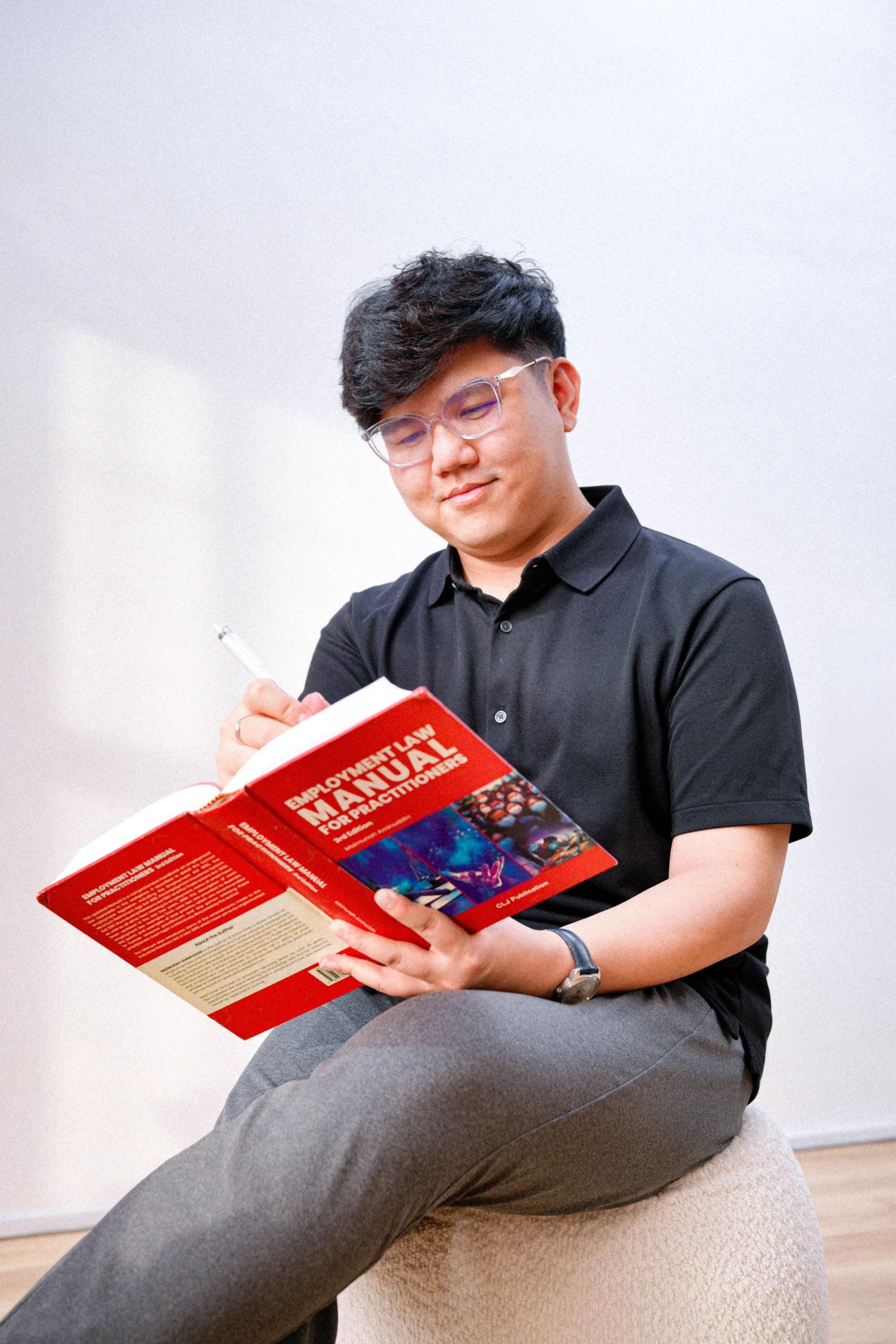 A man sitting on a stool, writing in a book titled 'Manual for Professionals' with a pen.