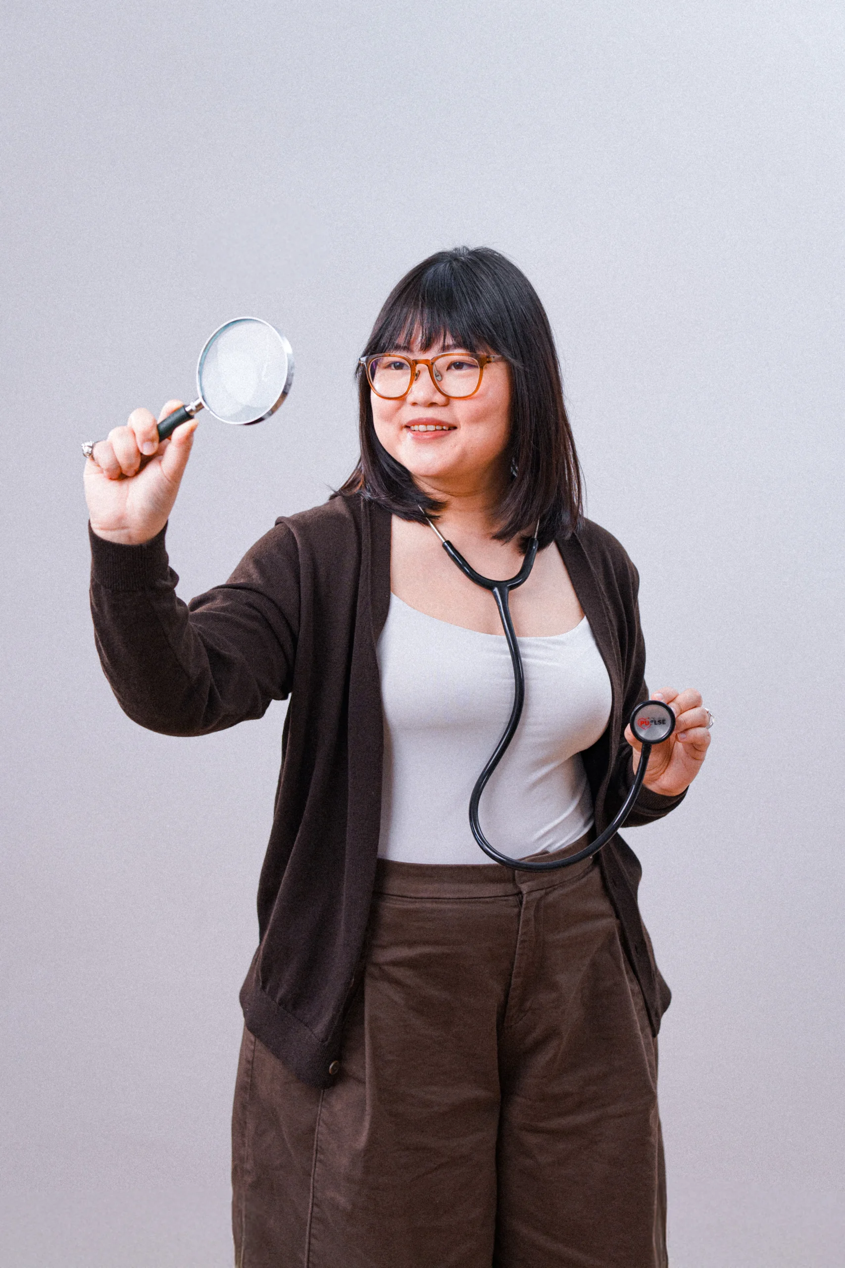 A woman holding a magnifying glass and wearing a stethoscope, looking inquisitive and smiling.